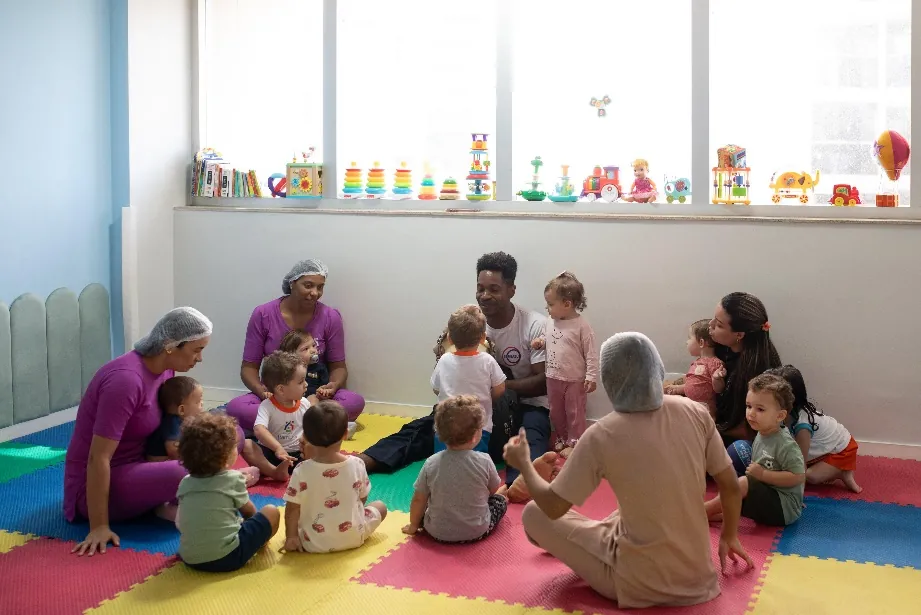 Crianças e educadoras sentadas em roda sobre tapete colorido